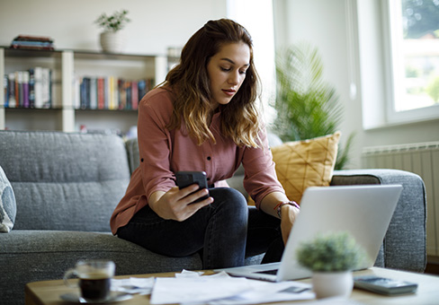 Woman sitting on couch with phone in hand and on laptop